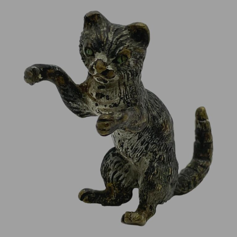 Main photo of small antique bronze cat displayed upright on its backside and facing towards the bottom left corner. Its front right paw is raised as if pawing