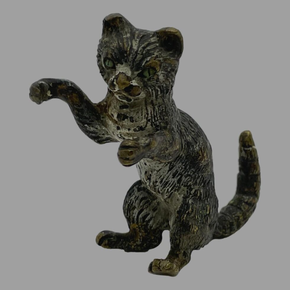 Main photo of small antique bronze cat displayed upright on its backside and facing towards the bottom left corner. Its front right paw is raised as if pawing
