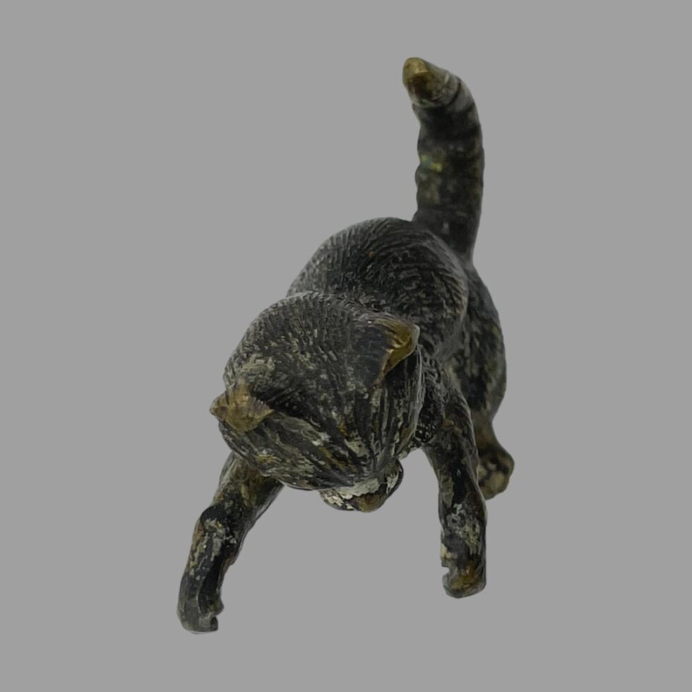 Photo of small antique bronze cat seen on all fours with its head in the centre foreground. The tail is upright in the back.