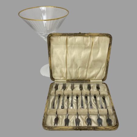 Main photo of cased cocktail forks set displayed inside their original fitted case with the lid leaning against a lovely cocktail glass (for display only) The case interior is in good used condition with only minor staining and some foxing. The forks are neatly laid out tail to tail with the 2 pronged ends on the outer ends. The clasp is unfortunately missing the closure.