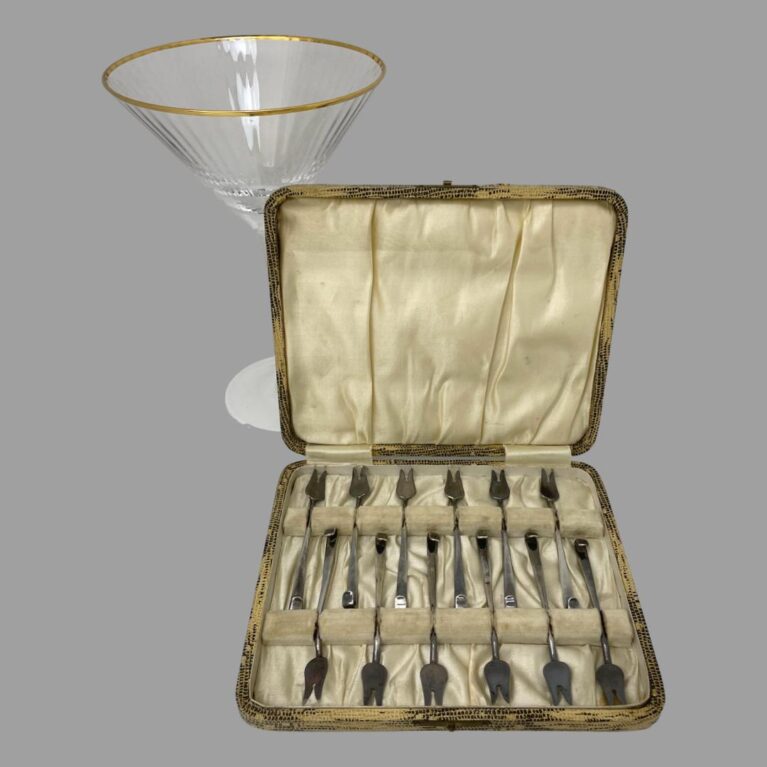 Main photo of cased cocktail forks set displayed inside their original fitted case with the lid leaning against a lovely cocktail glass (for display only) The case interior is in good used condition with only minor staining and some foxing. The forks are neatly laid out tail to tail with the 2 pronged ends on the outer ends. The clasp is unfortunately missing the closure.