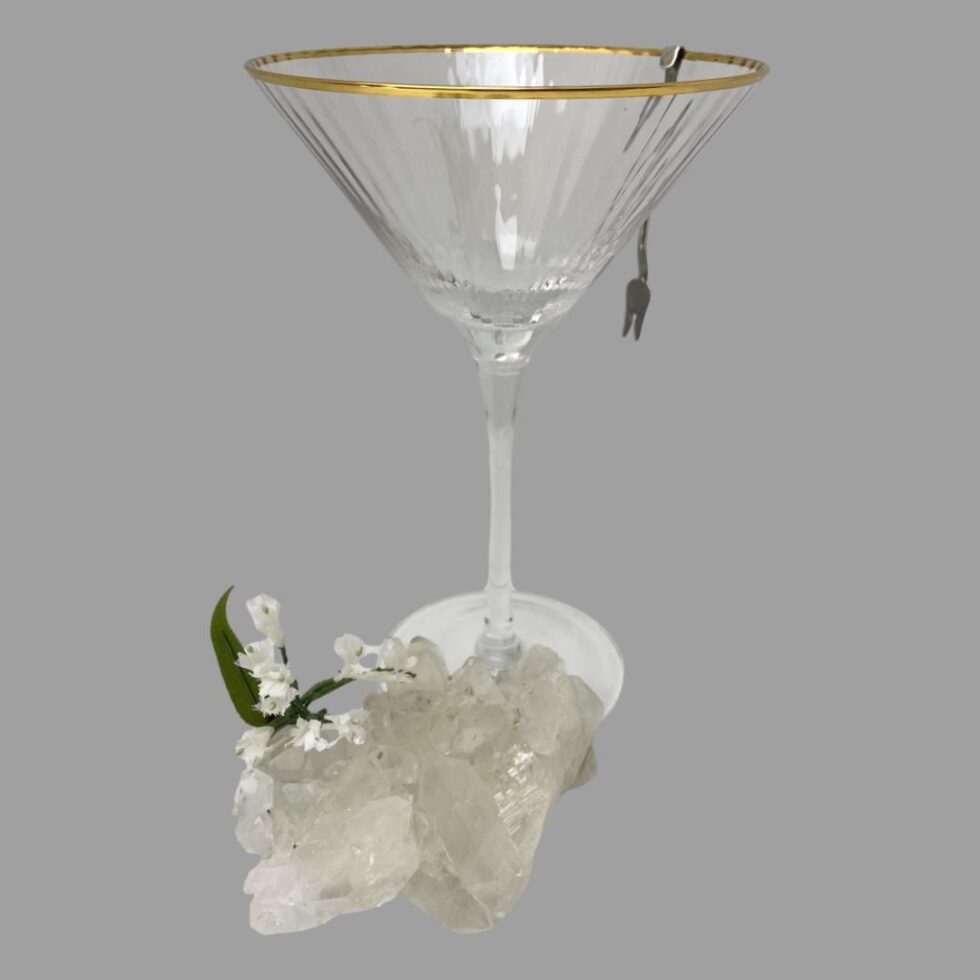 Photo of one of the forks hanging off the side of a cocktail glass by its hook. There is a small rock of quartz crystal in the foreground with a small spray of white flowers on top. Just a display detail.