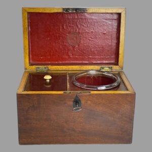 Main photo of small antique tea caddy seen from the front with lid open. The glass mixing bowl is in its slot on the right and the caddy on the left with its lid on. The inside of the lid and glass mixing slot is a bright red. The box is plain and straight sided with a metal ring to the centre of the lid.