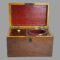 Main photo of small antique tea caddy seen from the front with lid open. The glass mixing bowl is in its slot on the right and the caddy on the left with its lid on. The inside of the lid and glass mixing slot is a bright red. The box is plain and straight sided with a metal ring to the centre of the lid.