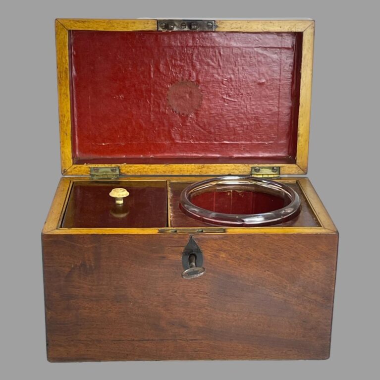 Main photo of small antique tea caddy seen from the front with lid open. The glass mixing bowl is in its slot on the right and the caddy on the left with its lid on. The inside of the lid and glass mixing slot is a bright red. The box is plain and straight sided with a metal ring to the centre of the lid.