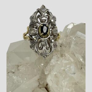 Main photo of Edwardian sapphire diamonds ring displayed on top of a rock of natural quartz crystal with the ring front forward facing. The oval cut sapphire in the centre has its own setting in gold with 4 teeth holding it in place. The elaborate silver front is added to the cover the rest of the rings face with the 10 old rose cut diamonds spaced out evenly throughout. The ring is vertically long with pointed top and bottom. The back of the elaborate face is yellow gold so the silver work at the front is like a shield or mask. Nice split band at the shoulders add an elegant touch,