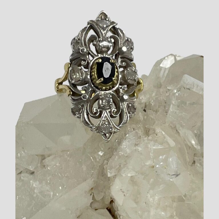 Main photo of Edwardian sapphire diamonds ring displayed on top of a rock of natural quartz crystal with the ring front forward facing. The oval cut sapphire in the centre has its own setting in gold with 4 teeth holding it in place. The elaborate silver front is added to the cover the rest of the rings face with the 10 old rose cut diamonds spaced out evenly throughout. The ring is vertically long with pointed top and bottom. The back of the elaborate face is yellow gold so the silver work at the front is like a shield or mask. Nice split band at the shoulders add an elegant touch,