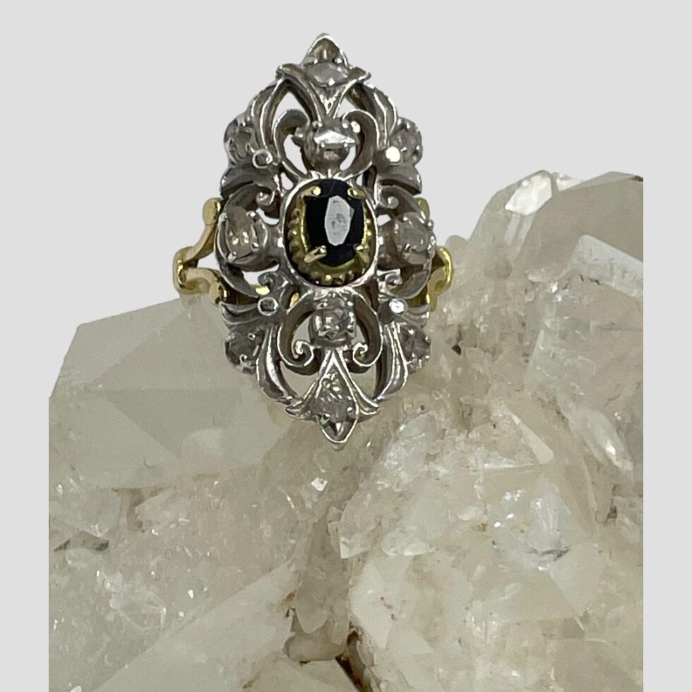 Main photo of Edwardian sapphire diamonds ring displayed on top of a rock of natural quartz crystal with the ring front forward facing. The oval cut sapphire in the centre has its own setting in gold with 4 teeth holding it in place. The elaborate silver front is added to the cover the rest of the rings face with the 10 old rose cut diamonds spaced out evenly throughout. The ring is vertically long with pointed top and bottom. The back of the elaborate face is yellow gold so the silver work at the front is like a shield or mask. Nice split band at the shoulders add an elegant touch,