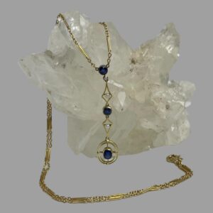 Main photo of antique gold &sapphire necklace displayed draped over a rock of natural quartz crystal with the attached pendant at the centre. The chain is over the quartz and appears from behind and laid across the front of the quartz with the clasp ending on the right. The dangling pendant is attached to the ends of the chain and has a round cut sapphire at the top with a seed pearl immediately below. This is followed by a diamond shaped frame in gold with a seed pearl at the centre. Another round cut sapphire follows this with another diamond shaped gold frame with central seed pearl and finally a double lined gold oval frame with the largest of the sapphires in an oval cut at the centre.