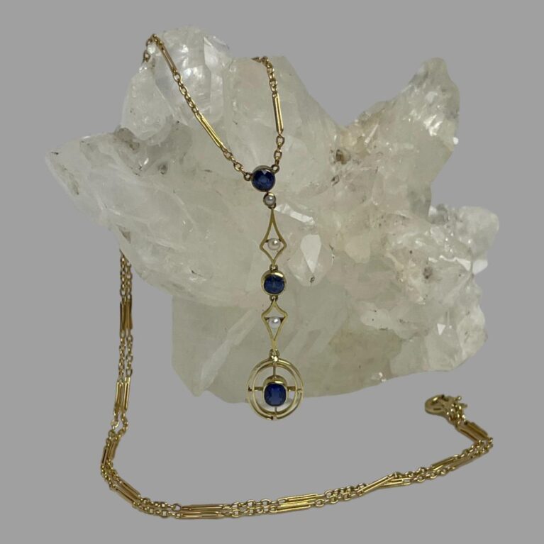 Main photo of antique gold &sapphire necklace displayed draped over a rock of natural quartz crystal with the attached pendant at the centre. The chain is over the quartz and appears from behind and laid across the front of the quartz with the clasp ending on the right. The dangling pendant is attached to the ends of the chain and has a round cut sapphire at the top with a seed pearl immediately below. This is followed by a diamond shaped frame in gold with a seed pearl at the centre. Another round cut sapphire follows this with another diamond shaped gold frame with central seed pearl and finally a double lined gold oval frame with the largest of the sapphires in an oval cut at the centre.