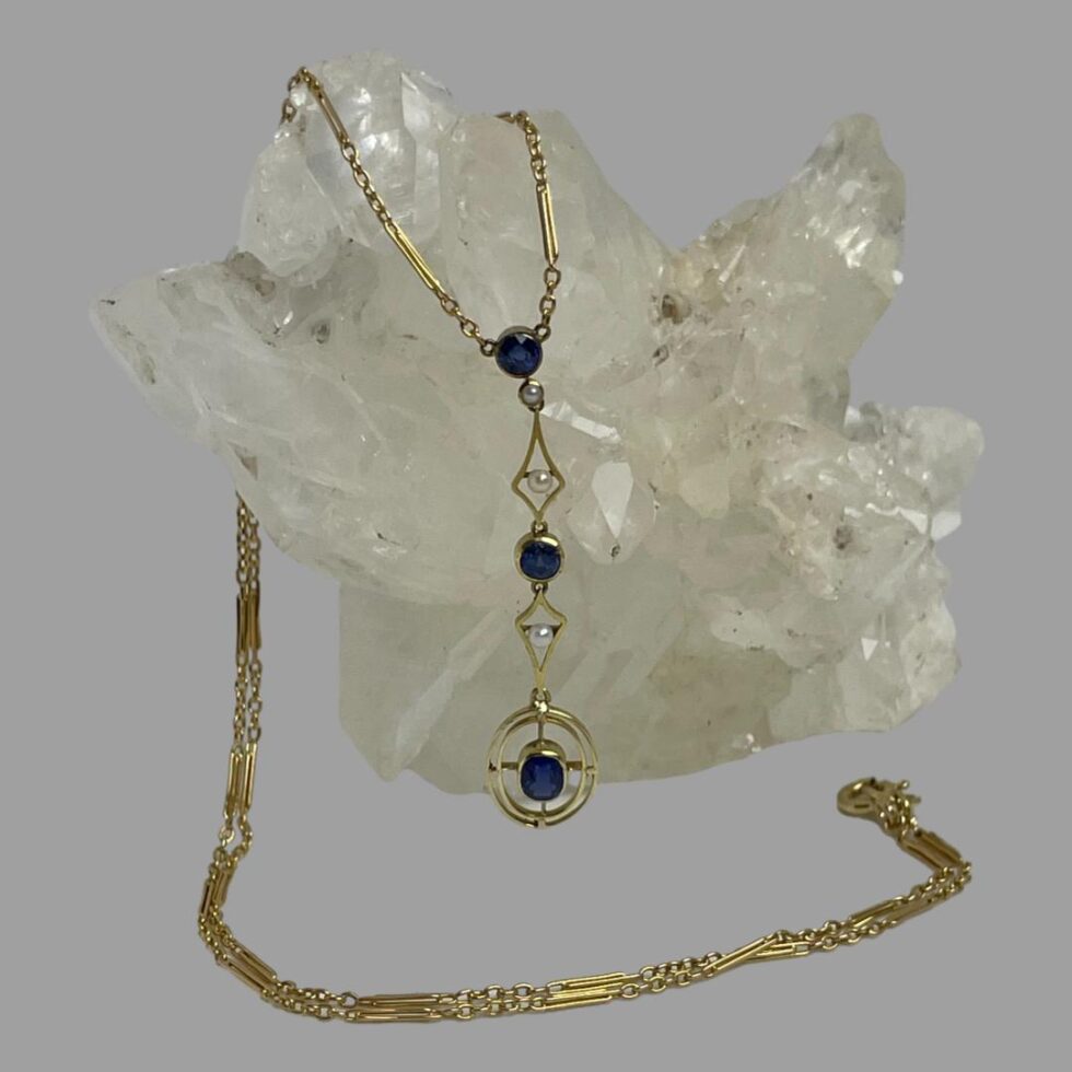 Main photo of antique gold &sapphire necklace displayed draped over a rock of natural quartz crystal with the attached pendant at the centre. The chain is over the quartz and appears from behind and laid across the front of the quartz with the clasp ending on the right. The dangling pendant is attached to the ends of the chain and has a round cut sapphire at the top with a seed pearl immediately below. This is followed by a diamond shaped frame in gold with a seed pearl at the centre. Another round cut sapphire follows this with another diamond shaped gold frame with central seed pearl and finally a double lined gold oval frame with the largest of the sapphires in an oval cut at the centre.