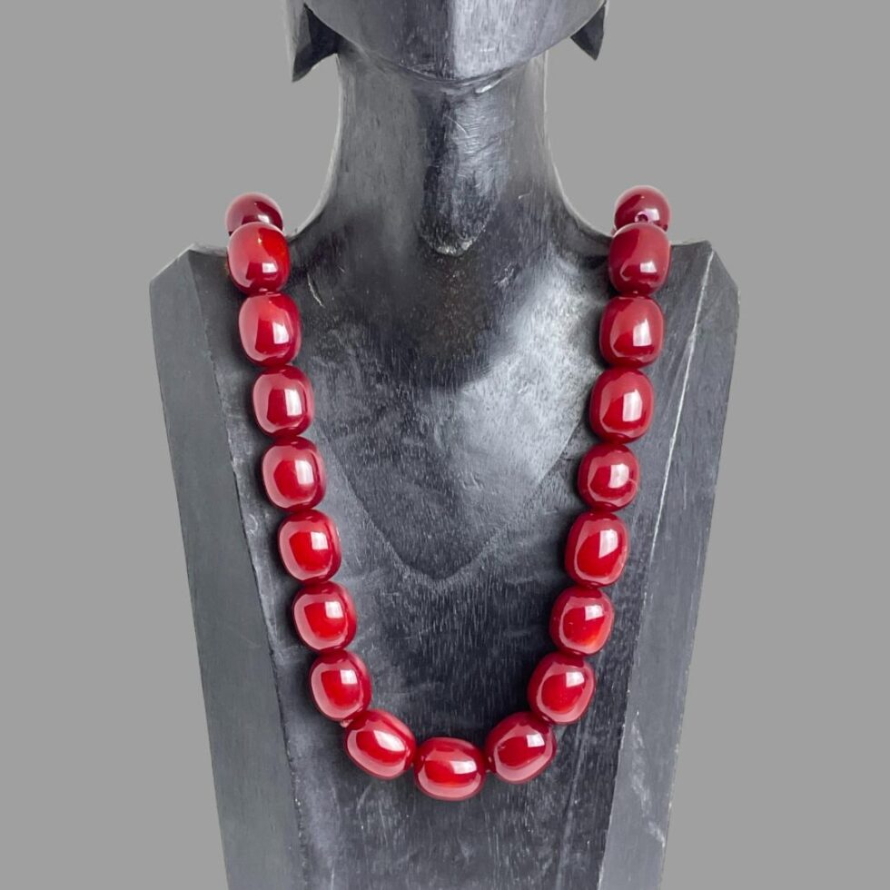 Main photo of cherry bakelite amber necklace displayed on a black wooden display stand and forward facing. The beads of this necklace are large barrel shaped and the colour is absolutely cherry red!