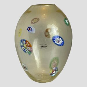 Main photo of 1970s Murano glass vase seen from an eye level showing the side with the original sticker label on. The vase has an almost egg like shape with the pointier end of 'the egg' at the base. The murrine areas are scattered around the vase and all are different in size and colour. The main body of the vase is a slightly milky glass with gold dust flecks inside.