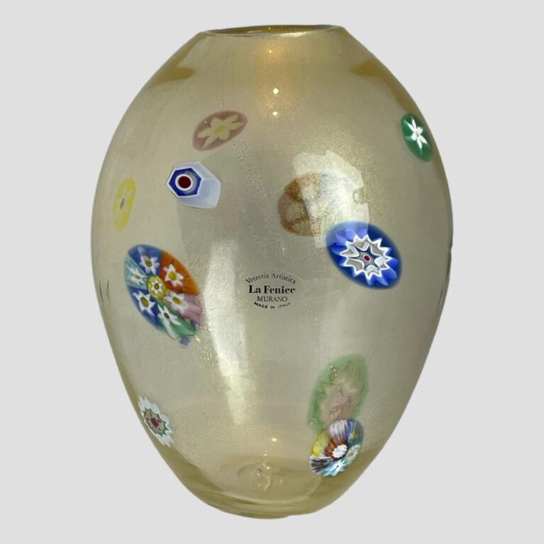 Main photo of 1970s Murano glass vase seen from an eye level showing the side with the original sticker label on. The vase has an almost egg like shape with the pointier end of 'the egg' at the base. The murrine areas are scattered around the vase and all are different in size and colour. The main body of the vase is a slightly milky glass with gold dust flecks inside.