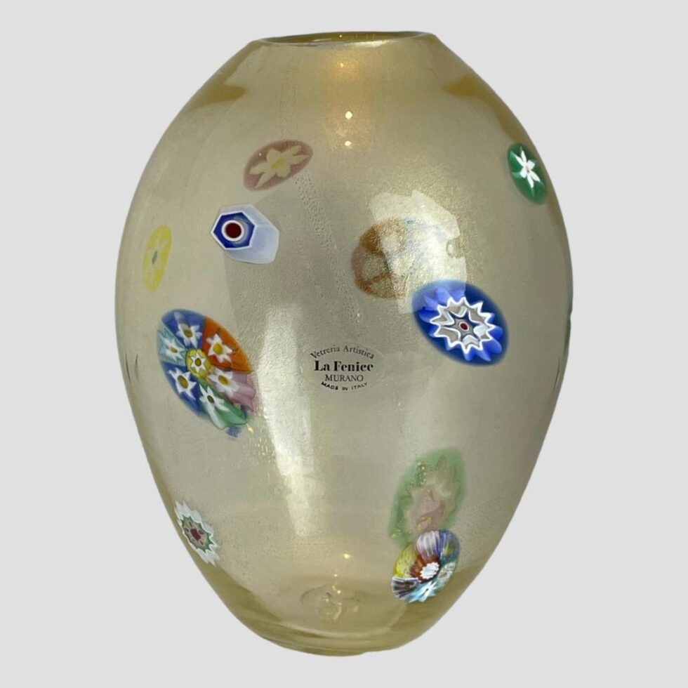 Main photo of 1970s Murano glass vase seen from an eye level showing the side with the original sticker label on. The vase has an almost egg like shape with the pointier end of 'the egg' at the base. The murrine areas are scattered around the vase and all are different in size and colour. The main body of the vase is a slightly milky glass with gold dust flecks inside.