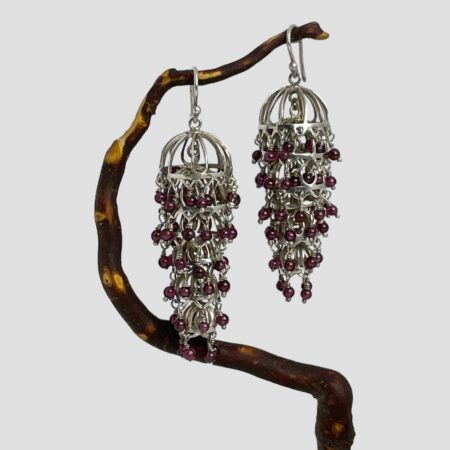 Main photo of silver garnet chandelier earrings displayed hanging from a twig of cherry tree. The twig bends upward so the earrings are not on the level with left one slightly lower than the right. The articulated design means these earrings move with your movement and always the right way round! The earrings have been called chandelier because of the way they hang but they could also be described as flapper dress and would actually make great accessories for a 1920s them look. The garnets are a beautiful shade of deep pink with a purple hew - like red grapes but tinier!