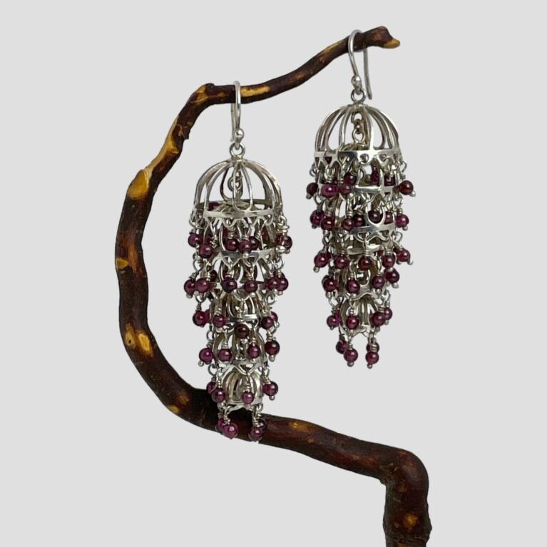 Main photo of silver garnet chandelier earrings displayed hanging from a twig of cherry tree. The twig bends upward so the earrings are not on the level with left one slightly lower than the right. The articulated design means these earrings move with your movement and always the right way round! The earrings have been called chandelier because of the way they hang but they could also be described as flapper dress and would actually make great accessories for a 1920s them look. The garnets are a beautiful shade of deep pink with a purple hew - like red grapes but tinier!