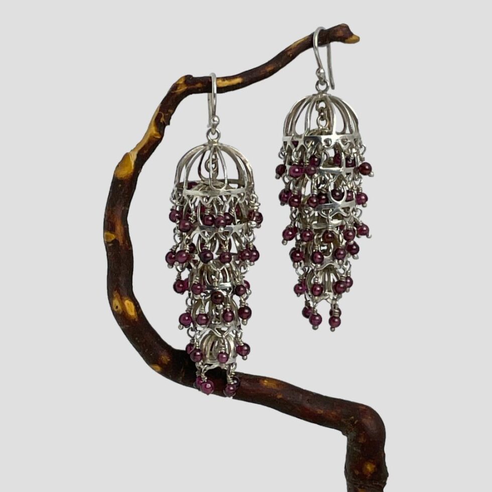 Main photo of silver garnet chandelier earrings displayed hanging from a twig of cherry tree. The twig bends upward so the earrings are not on the level with left one slightly lower than the right. The articulated design means these earrings move with your movement and always the right way round! The earrings have been called chandelier because of the way they hang but they could also be described as flapper dress and would actually make great accessories for a 1920s them look. The garnets are a beautiful shade of deep pink with a purple hew - like red grapes but tinier!