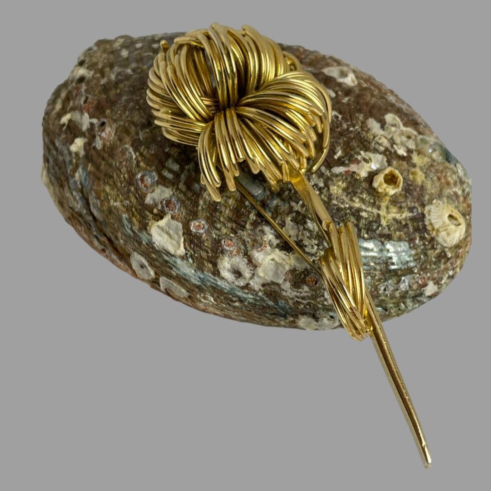 Main photo of signed Dior flower brooch displayed with flower head leaning on an upturned shell. The stem of the flower is quite long and rests on the flat surface. The flower head and leaves are constructed from strands of gilt metal almost hairlike quality