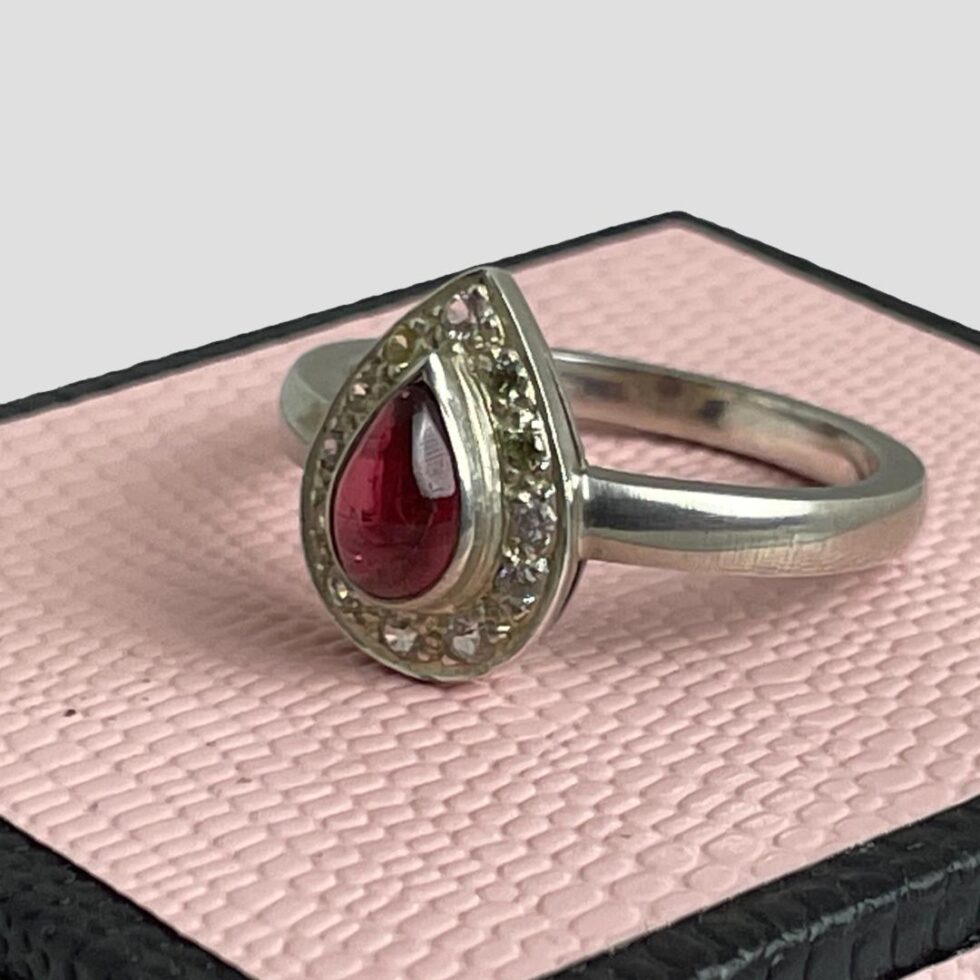 Photo of tourmaline & sapphire ring on top of its ring box with the ring front facing towards the bottom left. From this angle you can see that the tourmaline is set slightly raised from the white sapphire frame which is set back.