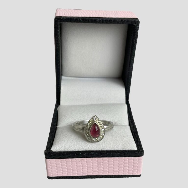 Main photo of tourmaline & sapphire ring displayed inside its ring box which has a baby pink colour exterior with black beading. The ring is forward facing with the pointed end at the top.