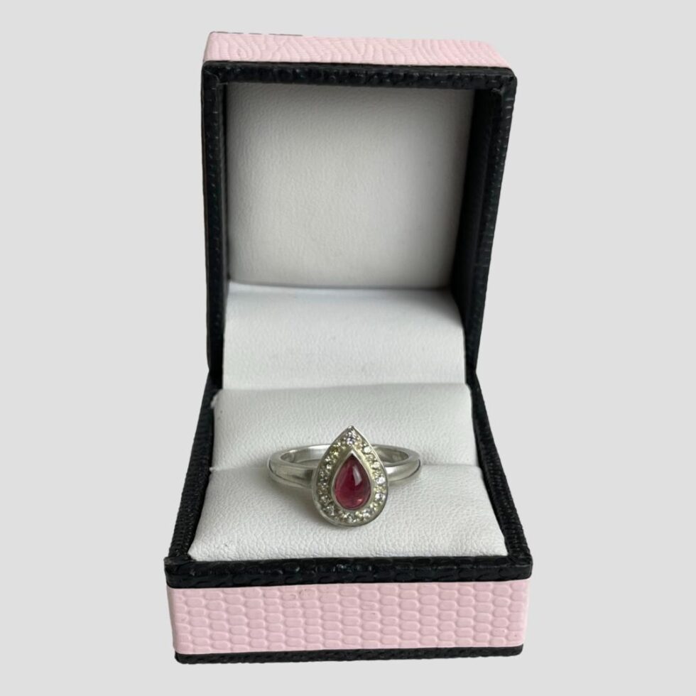 Main photo of tourmaline & sapphire ring displayed inside its ring box which has a baby pink colour exterior with black beading. The ring is forward facing with the pointed end at the top.
