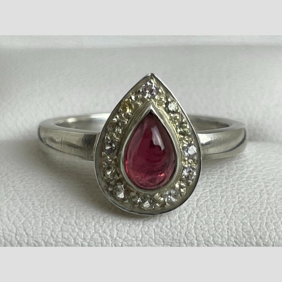 A close up photo of the tourmaline and sapphire ring front. The pointed end is at the top but the ring can be worn either way up as preferred. The pear or tear drop shape of the cerise tourmaline is mirrored in the ring design with the white sapphires set in a pear/teardrop shaped frame and set on sterling silver.