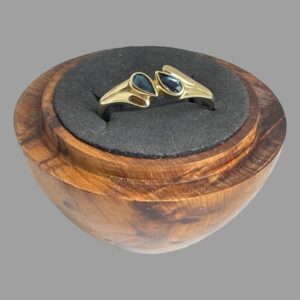 Main photo of gold blue gemstone ring displayed inside a wooden ring display stand with black interior. The ring front is forward facing and slightly upward showing the 2 gemstones. The pear or tear drop cut stones are set top to tail diagonally to the left and right of the band and held at the middle with a short gold divider. The ring shoulders are wider than the rest of the ring band to accommodate the diagonally set stones. The shoulders have 2 parts ; a shorter one with the wider end of the stone set in and a longer almost water droplet like extended piece which runs along the side of one pear shaped stone with the pointed ends at the centre. The ring appears like it has a split band but is solidly set as a whole ring.