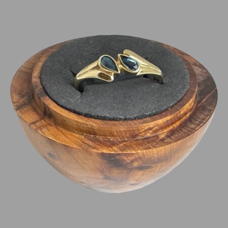 Main photo of gold blue gemstone ring displayed inside a wooden ring display stand with black interior. The ring front is forward facing and slightly upward showing the 2 gemstones. The pear or tear drop cut stones are set top to tail diagonally to the left and right of the band and held at the middle with a short gold divider. The ring shoulders are wider than the rest of the ring band to accommodate the diagonally set stones. The shoulders have 2 parts ; a shorter one with the wider end of the stone set in and a longer almost water droplet like extended piece which runs along the side of one pear shaped stone with the pointed ends at the centre. The ring appears like it has a split band but is solidly set as a whole ring.