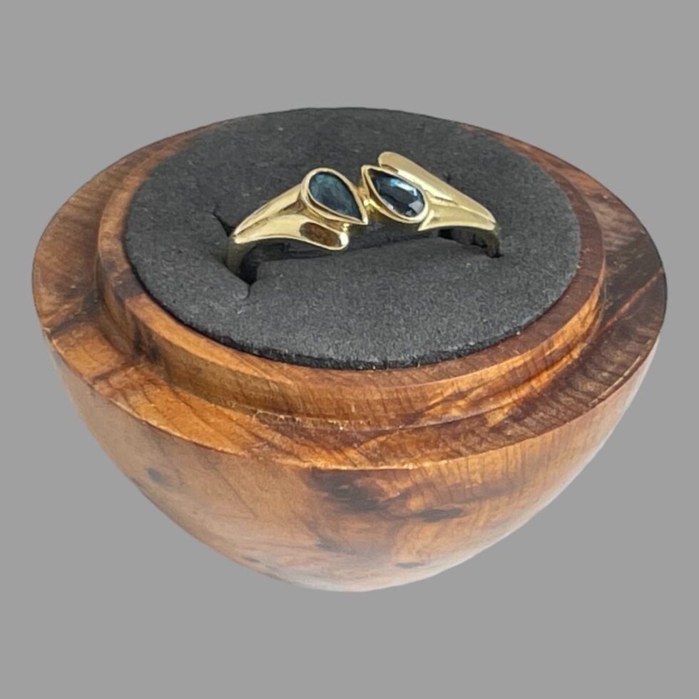 Main photo of gold blue gemstone ring displayed inside a wooden ring display stand with black interior. The ring front is forward facing and slightly upward showing the 2 gemstones. The pear or tear drop cut stones are set top to tail diagonally to the left and right of the band and held at the middle with a short gold divider. The ring shoulders are wider than the rest of the ring band to accommodate the diagonally set stones. The shoulders have 2 parts ; a shorter one with the wider end of the stone set in and a longer almost water droplet like extended piece which runs along the side of one pear shaped stone with the pointed ends at the centre. The ring appears like it has a split band but is solidly set as a whole ring.