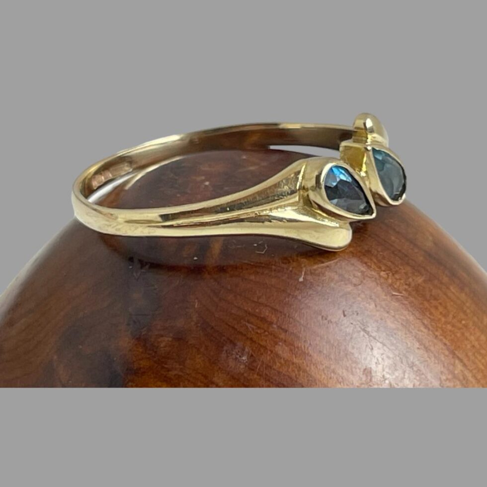 Photo of gold blue gemstone ring displayed on a wooden stand with the ring front facing right and showing the unusual shoulder design.