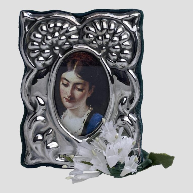 Main photo of small silver fronted frame displayed upright and forward facing. The design of the silver front is quite unusual with what could be 2 chrysanthemums or dandelion seed heads seen from a side angle to the top left and right of frame. The lower part is possibly foliage but with no definite pattern. From a distance, there is an owl like look. The photo display area is a longish thin oval and there is a picture of a thoughtful looking young lady with eyes downward and wearing a slight smile. There is also a small spray of artificial small white flowers laid at the bottom obscuring the hallmark on the right with just part of the makers initials on display at left.
