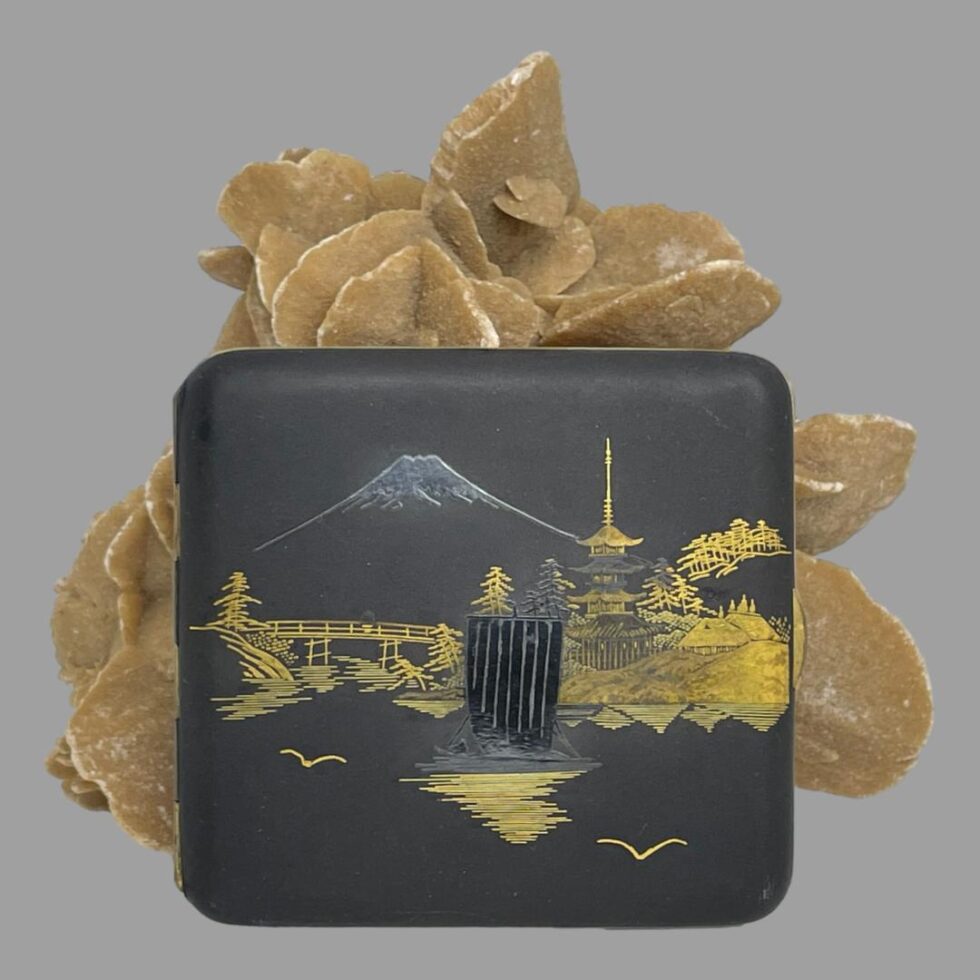 Main photo of Japanese damascene cigarette case displayed upright leaning against a rock of sand rose with the decorative side on display. The image is of Mount Fuji in the background with silver top and a boat with tall sails also in silver in the centre. The rest of the scene is in 24 karat gold inlay showing a pagoda in the background on right, with temple and old thatch houses and trees. On the left is a wooden bridge, some more trees. In very good condition for its age with no dents and only minor signs of wear namely scuff marks to the blackened metal base.