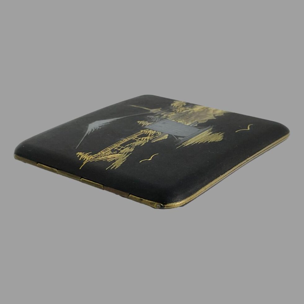 Photo of Japanese damascene cigarette case laid on a flat surface with the bottom left corner of the case in the centre foreground. This is to show the depth of the case and the slight curve for ease of fitting in pocket. At this angle you can see the silver of the tip of Mount Fuji and the sails of the boat.