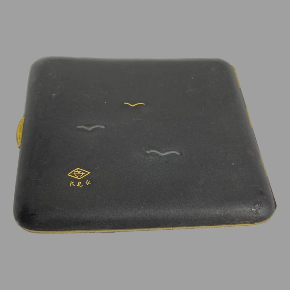 Photo of the back of the damascene cigarette case which is laid face down. The curve of the case design is very clear in this photo. The sparse 3 bird design and stylised signature is to the bottom left.
