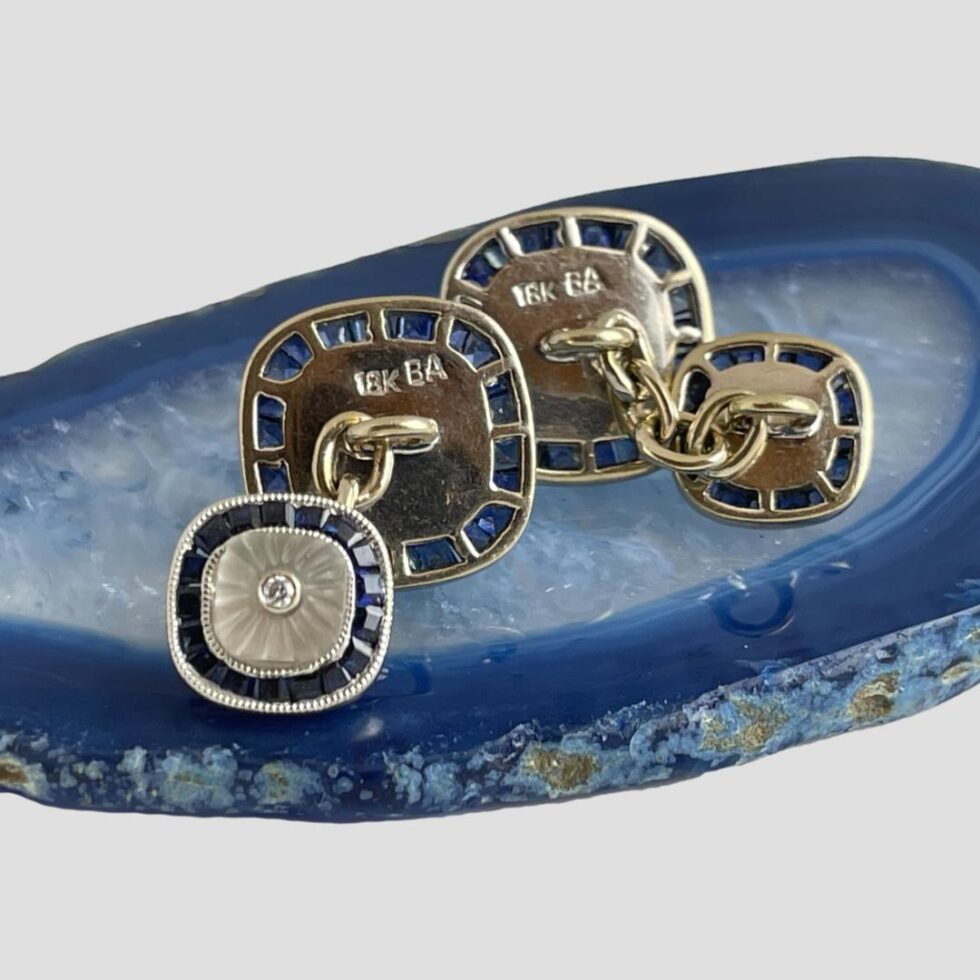Photo of the white gold sapphire cufflinks still displayed on a small slice of blue agate this time showing the back and the hallmark. The hallmarks are on the back of the larger squares which are both faced away from camera. The lefthand cufflink has the smaller square forward facing and the righthand one is showing the back of both larger and small.