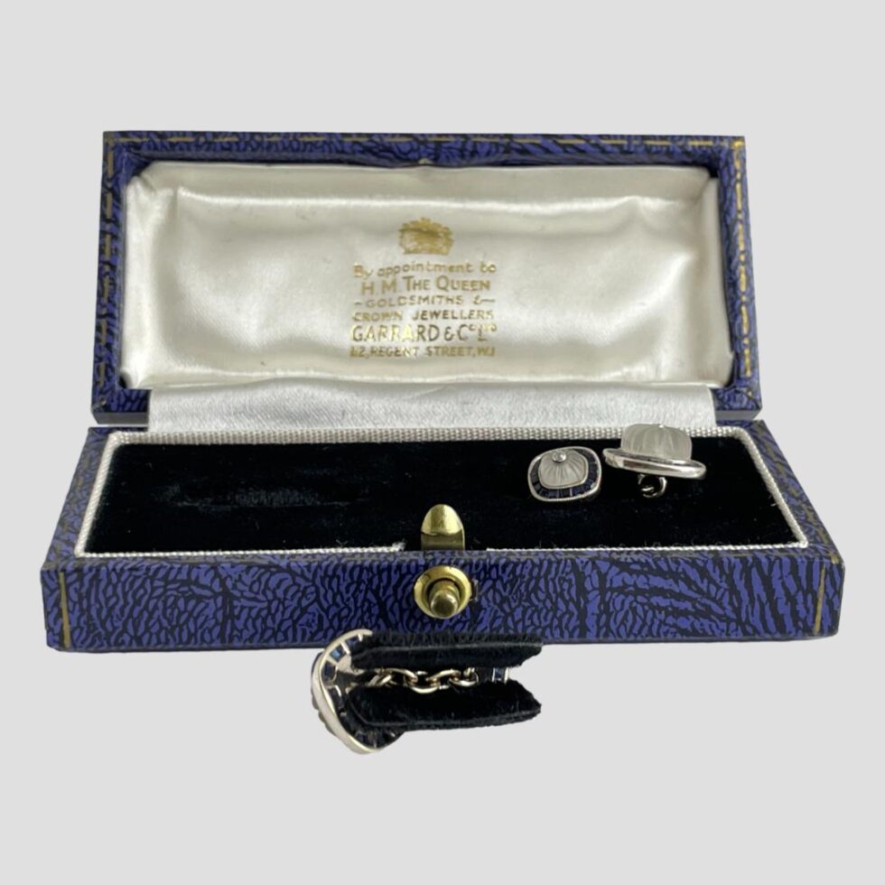 Photo of white gold sapphire cufflinks with one still inside the box and other one removed and placed in front of box. The cufflink outside is still sandwiched in to the velvet holder (a small folded piece of velvet covered card. The cufflinks chain runs along the folded part and the whole thing neatly fits into the slot inside the box.