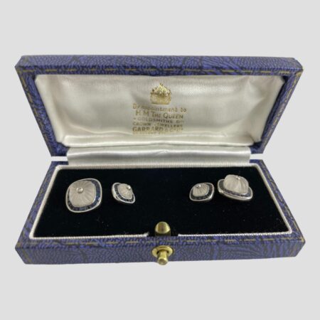 Main photo of white gold sapphire cufflinks displayed inside the Garrard & Co cufflinks box. The open box is forward facing with the cufflinks securely in place to the left and right. The box has a blue exterior, an almost black blue velvet inner and white silk lined inside the lid with Garrard & Co details in gilt. The larger of the link squares are on the outer extremes and the smaller squares inside. The cufflinks design - all uniform except size - square shaped but with rounded edges. A carved rock crystal sits in the centre of each also round-cornered squares. The carving in the crystal is lines emanating from the small round cut diamond at the centre. Around the perimeter is a frame of Channel set sapphires and all set in 18 karat white gold. Each area, the diamond, the rock crystal and the sapphires are separated with a white gold frame - the frame around the diamond is plain white gold whereas the frames around the rock crystal and sapphires have notches all the way around. An incredibly striking pair of cufflinks for the discerning man in your life!
