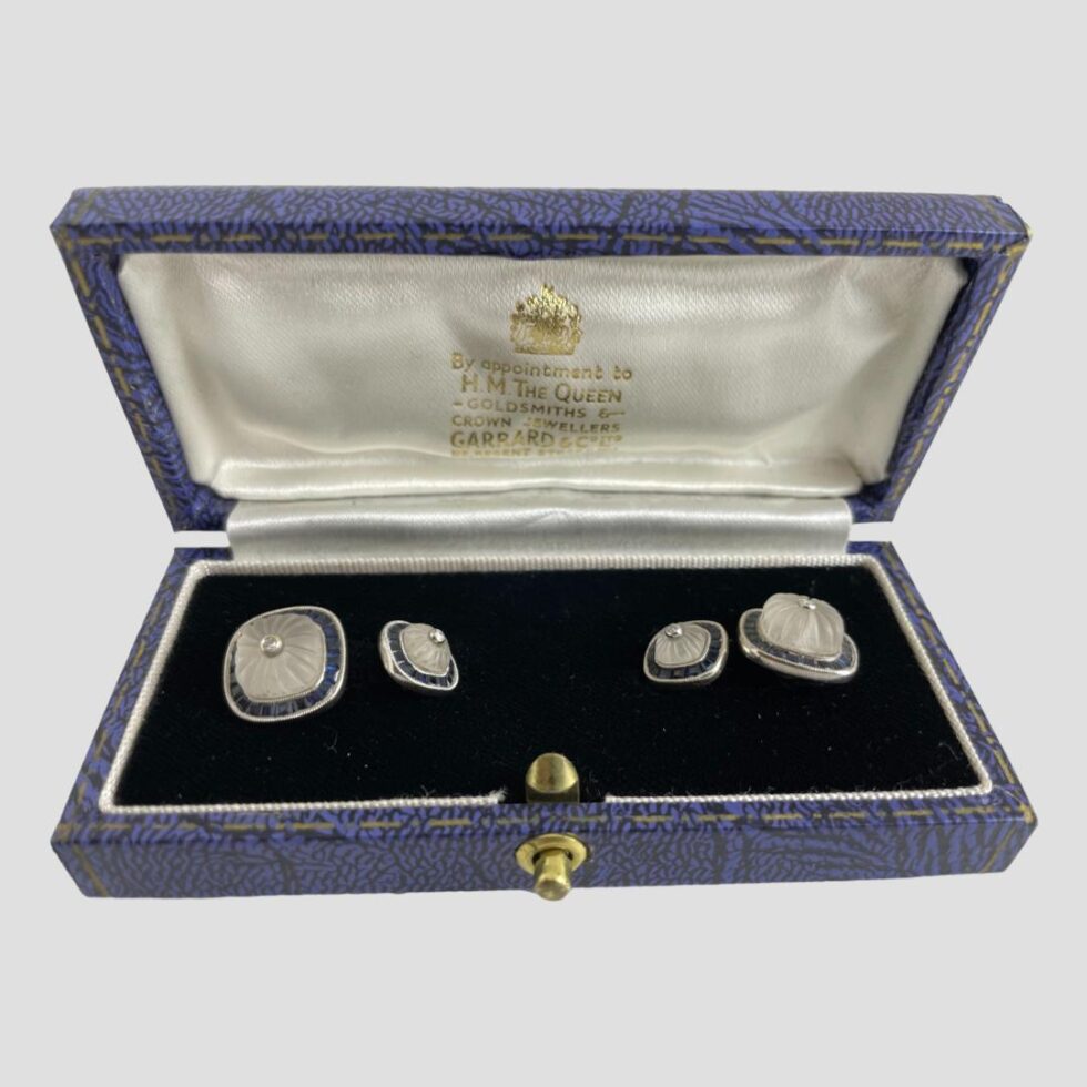 Main photo of white gold sapphire cufflinks displayed inside the Garrard & Co cufflinks box. The open box is forward facing with the cufflinks securely in place to the left and right. The box has a blue exterior, an almost black blue velvet inner and white silk lined inside the lid with Garrard & Co details in gilt. The larger of the link squares are on the outer extremes and the smaller squares inside. The cufflinks design - all uniform except size - square shaped but with rounded edges. A carved rock crystal sits in the centre of each also round-cornered squares. The carving in the crystal is lines emanating from the small round cut diamond at the centre. Around the perimeter is a frame of Channel set sapphires and all set in 18 karat white gold. Each area, the diamond, the rock crystal and the sapphires are separated with a white gold frame - the frame around the diamond is plain white gold whereas the frames around the rock crystal and sapphires have notches all the way around. An incredibly striking pair of cufflinks for the discerning man in your life!