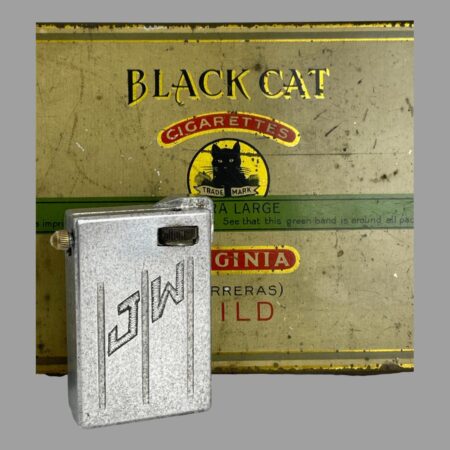 Main photo of trench art aluminium lighter displayed upright with the JW initialled side on display. The lift arm is shown closed and there is part of an old Black Cat brand cigarette tin behind for display purpose only. The flint wheel is visible to the top right corner of the lighter and this needs turning with a flick motion to create the spark. on the left edge, a brass tip is visible - this unscrews to add a new flint.