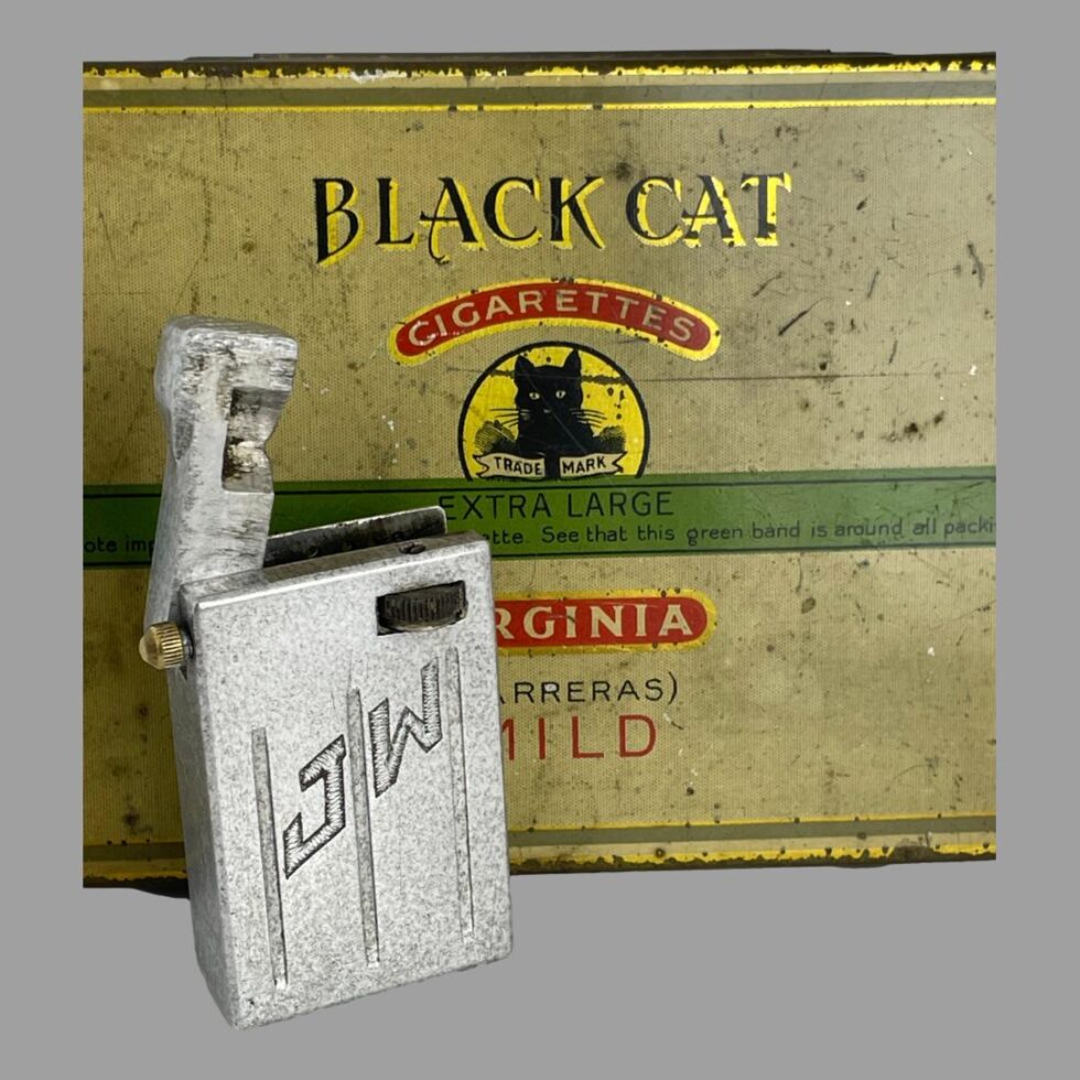 Photo of trench art aluminium lighter still displayed upright in front of an empty tin of Black Cat cigarettes this time with the lift arm open.