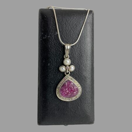 Main photo of ruby sapphire pendant &chain displayed hanging from the back of an old jewellery box. The chain has been wound round the box to secure the necklace and pendant in place. The main focus of this piece is without question the pendant. The carved ruby is a deep pink-almost cerise in colour and beautifully carved into a flower which covers the whole space. Around the floral ruby is a frame of silver with an outer edge of multiple coloured sapphires which include pink, blue, yellow, white and these enhance the floral ruby further. Above the ruby flower and multi coloured sapphire area is a bail separated section of 3 pearls (one at top and 2 below in a triangular formation) with a single pink sapphire at the centre. Above this is the generously sized large bail. The snake chain is suitable for this pendant and holds the weight well.