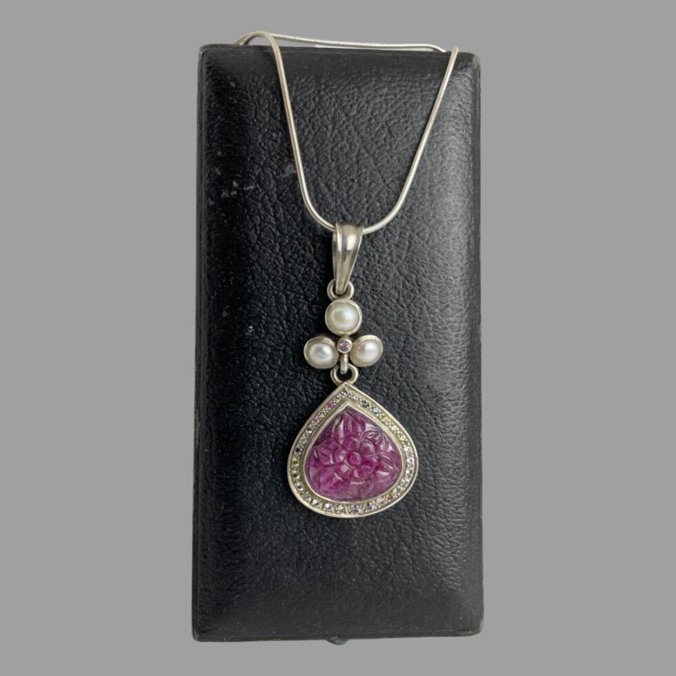 Main photo of ruby sapphire pendant &chain displayed hanging from the back of an old jewellery box. The chain has been wound round the box to secure the necklace and pendant in place. The main focus of this piece is without question the pendant. The carved ruby is a deep pink-almost cerise in colour and beautifully carved into a flower which covers the whole space. Around the floral ruby is a frame of silver with an outer edge of multiple coloured sapphires which include pink, blue, yellow, white and these enhance the floral ruby further. Above the ruby flower and multi coloured sapphire area is a bail separated section of 3 pearls (one at top and 2 below in a triangular formation) with a single pink sapphire at the centre. Above this is the generously sized large bail. The snake chain is suitable for this pendant and holds the weight well.