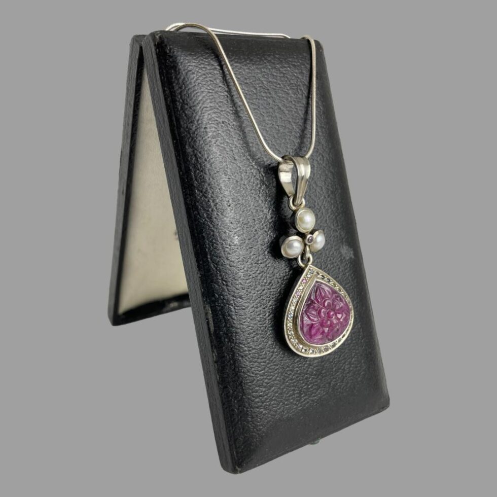 Photo of ruby sapphire pendant & chain still displayed on an old jewellery box this time facing to the right. You can see that the box is propping itself up with the inside of lid visible in the background.