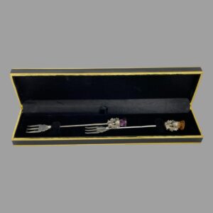 Main photo of the pair silver pickle forks displayed inside their case with the thistle ends at right and prongs at left. The fitted box holds the forks lengthways with a crossover section in the middle with one forks prongs and other forks thistle tip at centre. The box is not original to the forks but works very well.