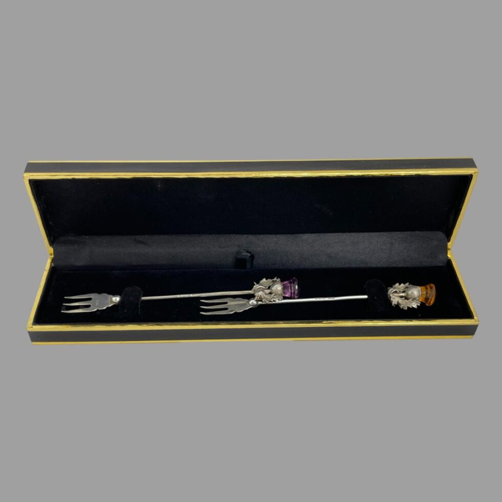 Main photo of the pair silver pickle forks displayed inside their case with the thistle ends at right and prongs at left. The fitted box holds the forks lengthways with a crossover section in the middle with one forks prongs and other forks thistle tip at centre. The box is not original to the forks but works very well.