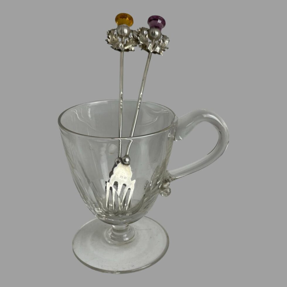 Photo of the 2 pickle forks displayed outside of the box and inside of a small antique glass. The thistle ends are sticking out at the top and the prong ends inside the glass.