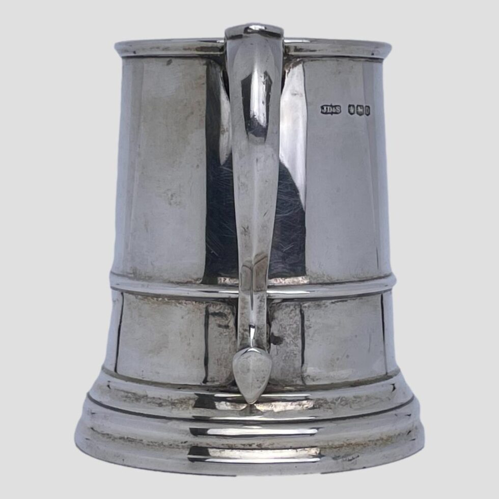 Another eye level photo of the silver half pint tankard this time with the handle in the centre. and bother sides of the tankard visible including hallmark on the right side.