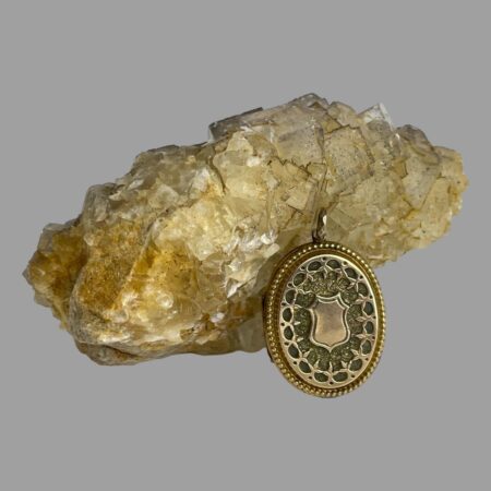 Main photo of antique gold overlay locket with the locket displayed leaning against a rock of river quartz The side of locket displayed is the front which has the larger empty cartouche. The design detail is very intricate with foliage style pattern around the cartouche and outer frame with brass 'jewels' (small balls) The locket is of a generous size.