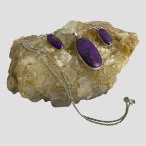 Main photo of purple Mojave turquoise set displayed on a rock of natural river quartz. The pendant on the necklace is at the centre with the chain swirled round to the left and the clasp area to the right in front of the rock. The earrings are flanked to the left and right of the pendant. The purple Mojave turquoise is like a patchwork of different purple shades. In addition to the purple shades, there are flecks of copper prominent mostly in one of the earrings on the left.